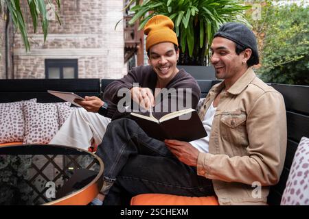 Un couple gay est assis sur un patio confortable, partageant un moment joyeux avec un livre. Entourés de plantes luxuriantes et d'oreillers moelleux, ils apprécient la compagnie de l'autre an Banque D'Images