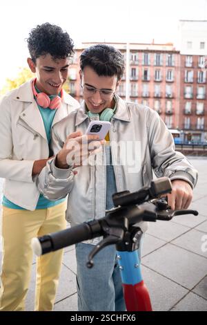 Un couple gay heureux, chacun portant des écouteurs, profite d'un moment de détente à l'extérieur tout en regardant un smartphone pour louer un scooter électrique. Banque D'Images