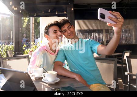 Un couple gay joyeux aime les moments ensoleillés, prenant un selfie ensemble dans un café en plein air. Ambiance détendue avec tasses à café et tablette numérique sur le Banque D'Images