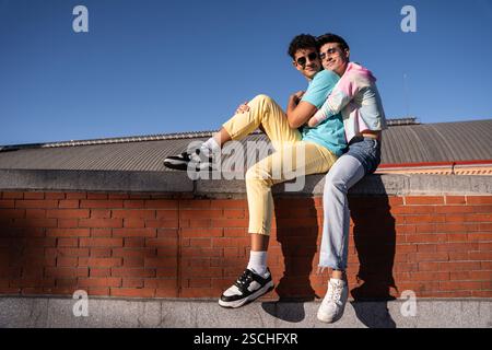 Un jeune couple gay joyeux est assis sur un mur de briques, embrassant et souriant sous un ciel bleu clair. Leur tenue décontractée et leurs lunettes de soleil reflètent un insouciant, s Banque D'Images