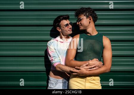 Un couple gay heureux partage une tendresse étreinte, avec un partenaire tenant l'autre par derrière. Ils se tiennent contre un mur ondulé vert, les deux portant s Banque D'Images
