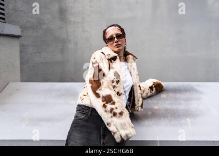 Femme élégante portant une veste de fourrure imprimée vache, des lunettes de soleil et du denim, appuyée contre une surface métallique dans un cadre urbain, incarnant la mode moderne et Banque D'Images