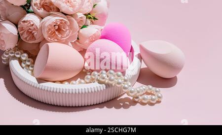 Diverses éponges d'oeufs de maquillage, un mélangeur de beauté, disposés au milieu de fausses perles et de roses roses sur un fond rose clair. Banque D'Images