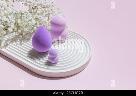 Différentes tailles d'éponges de maquillage Purple Beauty Blender disposées artistiquement sur une plaque texturée blanche, complétées par de délicates fleurs blanches sur un SAF Banque D'Images