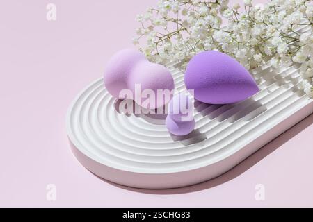 Différentes tailles et formes d'éponges de maquillage aux tons violets habilement mises en scène sur un plateau blanc strié, accompagnées de délicates fleurs blanches sur un dos rose Banque D'Images