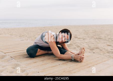Une femme pratique la pose de yoga Janu Sirsasana sur un tapis de bois à la plage, souriant à la caméra avec l'océan en arrière-plan pendant le coucher du soleil. Banque D'Images