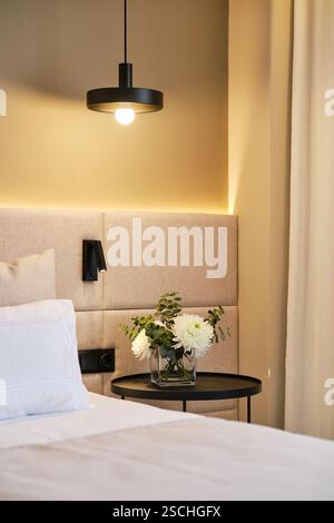 Un coin chambre d'hôtel confortable avec une lampe suspendue moderne, tête de lit rembourrée, et une table d'appoint avec un vase en verre de fleurs blanches, exsudant le confort et Banque D'Images