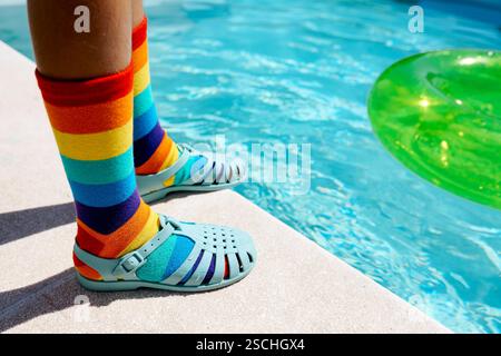 Une scène vibrante avec des chaussettes à rayures arc-en-ciel jumelées à des sandales bleues près d'une piscine lumineuse. Les chaussettes colorées et l'anneau gonflable vert ajoutent un Banque D'Images
