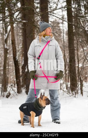 Une femme portant un manteau et un chapeau d'hiver promène un bouledogue français dans une forêt enneigée. Le chien porte une veste chaude, et une laisse rose vif ajoute une touche de col Banque D'Images