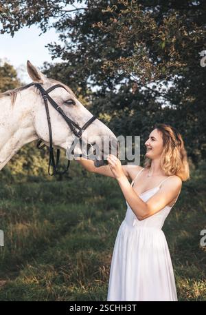 Une jeune femme souriante dans une robe blanche se tient en interaction ludique avec un cheval blanc. La scène se déroule dans un environnement extérieur luxuriant et verdoyant sous tr Banque D'Images
