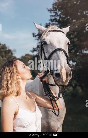 Une jeune femme en robe blanche partage un moment serein avec un cheval blanc en plein air. La lumière du soleil souligne leur lien, renforçant le paisible et natu Banque D'Images