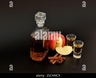 Liqueur de pomme rouge maison dans une carafe en cristal et deux verres sur fond noir, aux côtés de cannelle, d'anis et de morceaux de fruits mûrs. Gros plan. Banque D'Images