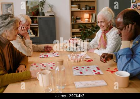 Plan à angle élevé d'une compagnie senior biraciale d'amis assis à la table en bois et jouant au bingo ensemble pendant que dans la maison de retraite Banque D'Images