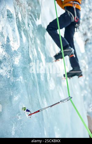 Gros plan mince quickdraw avec mousqueton rouge grimpant accroché les vis à glace annulaires vissées dans la glace d'un mur de glacier gelé contre un athlète grimpeur. Banque D'Images