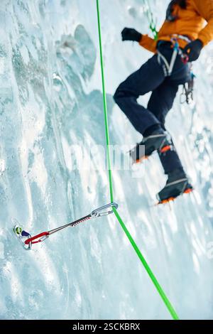 Gros plan mince quickdraw avec mousqueton rouge grimpant accroché les vis à glace annulaires vissées dans la glace d'un mur de glacier gelé contre un athlète grimpeur. Banque D'Images