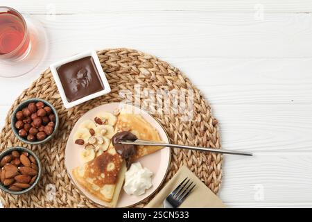 Délicieuses crêpes à la banane, pâte de chocolat, crème fouettée, noix et thé sur table en bois blanc, pose à plat Banque D'Images