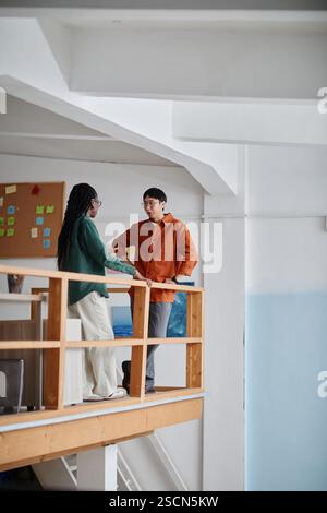 Deux collègues s'engageant dans la conversation dans un environnement de bureau contemporain avec des balustrades en bois et un tableau d'affichage en arrière-plan, démontrant la collaboration et le travail d'équipe Banque D'Images