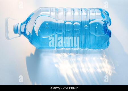 Grande bouteille en plastique remplie d'eau potable claire repose sur la surface blanche, reflétant la lumière du soleil et créant des motifs intéressants dans la zone environnante. Banque D'Images