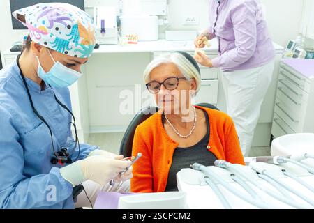 Dentiste portant un masque chirurgical et une loupe montrant une perceuse dentaire à un patient senior en clinique dentaire Banque D'Images