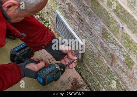 Un homme portant une veste rouge en utilisant une perceuse électrique avec un tournevis pour fixer un couvercle de maille de brique d'air à un mur. Le maillage empêche les souris d'entrer. Banque D'Images