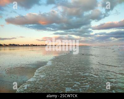 De douces vagues roulent sur la plage de sable tandis que les nuages colorés reflètent les teintes chaudes du coucher de soleil sur l'océan. Banque D'Images