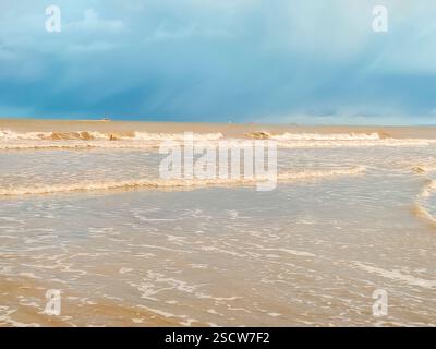 De douces vagues se battent contre la plage de sable tandis que des nuages sombres planent au-dessus de vous dans la lumière de la fin de l'après-midi. Banque D'Images