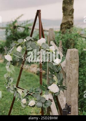 Un panneau de bienvenue floral est affiché sur un chevalet dans un lieu de mariage pittoresque, invitant les invités à célébrer l'occasion. Banque D'Images