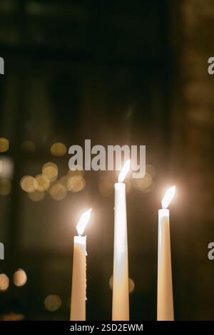 Trois grandes bougies blanches émettent une lumière douce dans une pièce légèrement éclairée, rehaussant l'ambiance chaleureuse de la soirée. Banque D'Images
