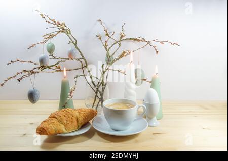 Pause café avec croissant et œuf dur, bouquet de branches de printemps avec des œufs de Pâques gris bleus suspendus et bougies allumées sur une table en bois agains Banque D'Images