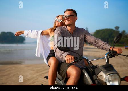 Un couple heureux fait de la moto sur une plage côtière pittoresque. L'homme conduit un scooter. La femme étend les bras en profitant du vent et du soleil. Ambiance tropicale de vacances. Parfait pour Banque D'Images