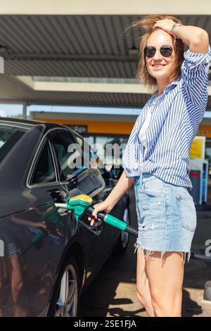 Portrait heureux souriant femme lunettes de soleil tenue décontractée ravitailler la station d'essence de voiture ensoleillée journée d'été. Femme confiante remplissant le réservoir de carburant automatique Banque D'Images
