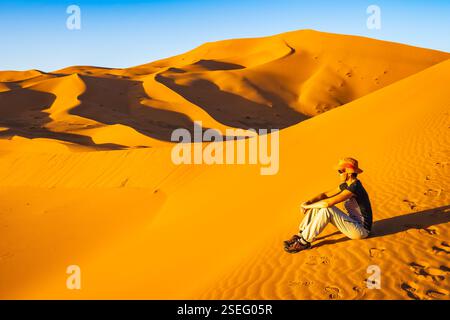 Jeune femme touriste avec chapeau assis sur la dune de sable à Erg Chebbi désert Sahara au coucher du soleil près de la ville de Merzouga, Maroc, Afrique du Nord Banque D'Images