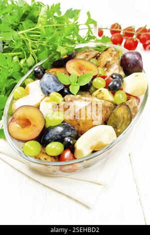 Poulet cuit au four avec tomates, les pommes, les prunes et les raisins dans un verre à rôtir sur une serviette légère, ail, persil et basilic sur fond blanc bois Banque D'Images