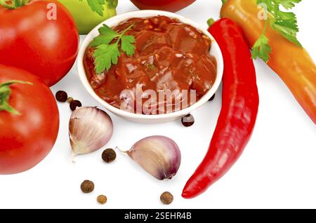 Ketchup dans une tasse blanche, tomates, deux piments rouges frais, ail, grains de poivre, moutarde, persil sur fond blanc Banque D'Images