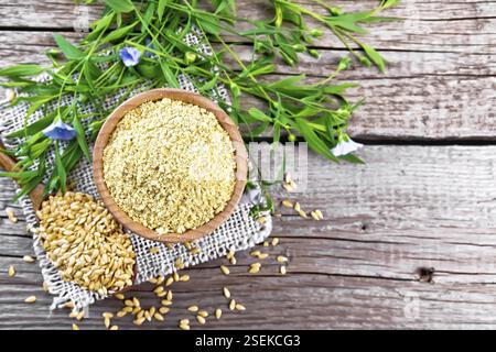 Son de lin dans un bol, graines dans une cuillère sur une serviette en toile de jute, feuilles de lin et fleurs sur le fond d'une vieille planche en bois d'en haut Banque D'Images