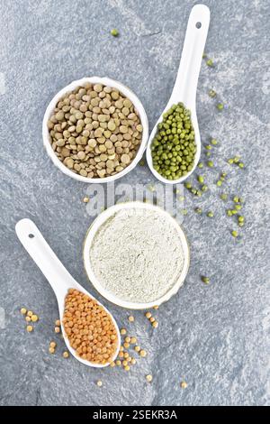 Farine de lentilles dans un bol, lentilles rouges, vertes dans des cuillères, brunes dans un bol, tiges de haricots avec des feuilles sur fond de table en pierre par le haut Banque D'Images