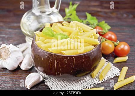 Pâtes de penne de farine de blé dans un bol de noix de coco sur le germe, tomates, ail, huile végétale dans un carafe et persil sur le backgro de bois foncé Banque D'Images