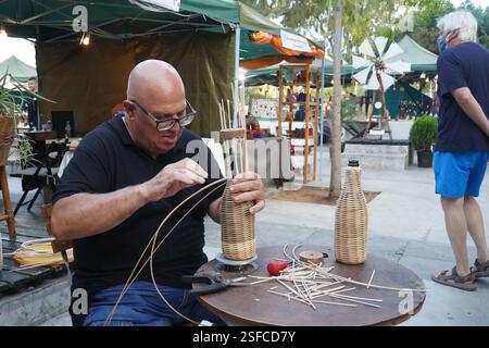 Un homme tisse une bouteille en osier, attachant des tiges de bois autour d'elle. Un espace de travail avec des matériaux prêts pour l'artisanat traditionnel mis sur table Banque D'Images