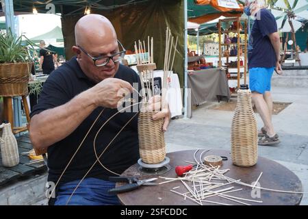 Un homme tisse une bouteille en osier, attachant des tiges de bois autour d'elle. Un espace de travail avec des matériaux prêts pour l'artisanat traditionnel mis sur table Banque D'Images