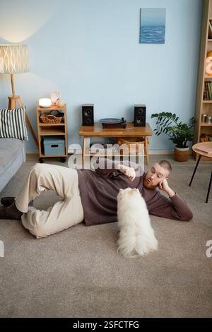 Personne allongée sur un tapis dans le salon tout en interagissant avec un chien moelleux, entourée d'un décor confortable et élégant avec une table tournante sur une table en bois et des étagères en arrière-plan Banque D'Images