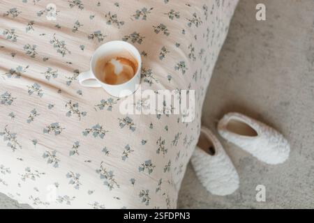 Vue aérienne d'une tasse à café vide sur un lit avec une couette fleurie et une paire de chaussons dans une chambre Banque D'Images