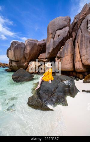 Une femme dans un chapeau à larges bords et une robe jaune est assise sur un grand rocher au bord de l'eau cristalline Banque D'Images