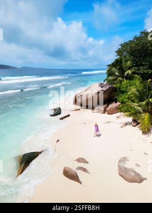Vue aérienne d'une femme en robe violette se promenant le long d'une plage immaculée sur la Digue, aux Seychelles, avec de grands rochers de granit et des palmiers luxuriants Banque D'Images