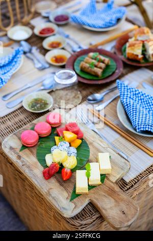 Un cadre de pique-nique magnifiquement aménagé avec une variété de desserts, y compris des macarons, des fruits tropicaux et des gâteaux, servis sur un plateau en bois Banque D'Images