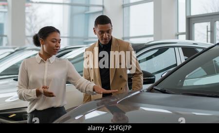Femme afro-américaine gestionnaire commerçant vendeur femme vendeur aider à consulter le client client masculin dans le salon automobile montrant la nouvelle voiture électrique moderne Banque D'Images