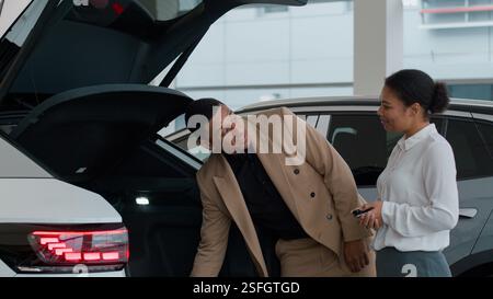 La femme de concessionnaire automobile démontre le client ouvrant automatiquement le coffre nouveau transport électrique moderne dans le salon de concession afro-américain d'acheteur d'homme Banque D'Images