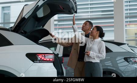 La femme de concessionnaire automobile démontre le client ouvrant automatiquement le coffre nouveau transport électrique moderne dans le salon de concession afro-américain d'acheteur d'homme Banque D'Images