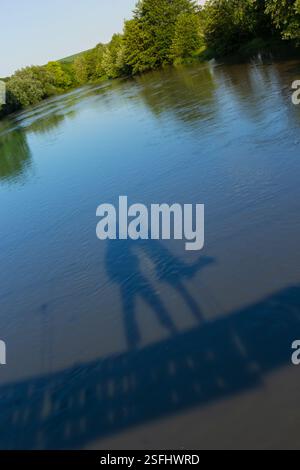 Deux silhouettes se tiennent près d'une rivière paisible, leurs ombres dansant à la surface de l'eau sous la lumière du soleil. Une végétation luxuriante entoure la scène, créa Banque D'Images