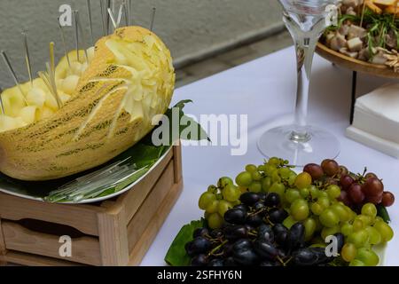 Une présentation de fruits magnifiquement agencée comprend un melon sculpté rempli de fruits en cubes, entouré de raisins assortis et d'un verre décoratif. Parfait fo Banque D'Images