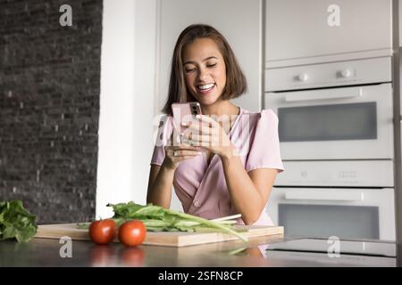 Jolie femme utilisant son smartphone se dresse dans la cuisine Banque D'Images